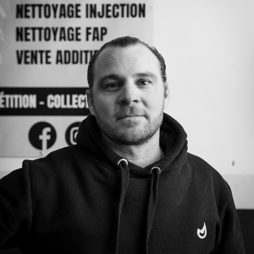 Spécialiste Nettoyage Injection FAP avec Services Additifs Homme barbu en sweat à capuche noir regardant l'objectif devant un panneau 'NETTOYAGE INJECTION FAP VENTE ADDITIFS'.