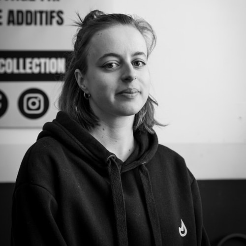 Portrait femme sweat noir collection additifs Portrait en noir et blanc d'une jeune femme portant un sweat à capuche noir avec un logo de flamme, devant une affiche mentionnant "ADDITIFS" et "COLLECTION".
