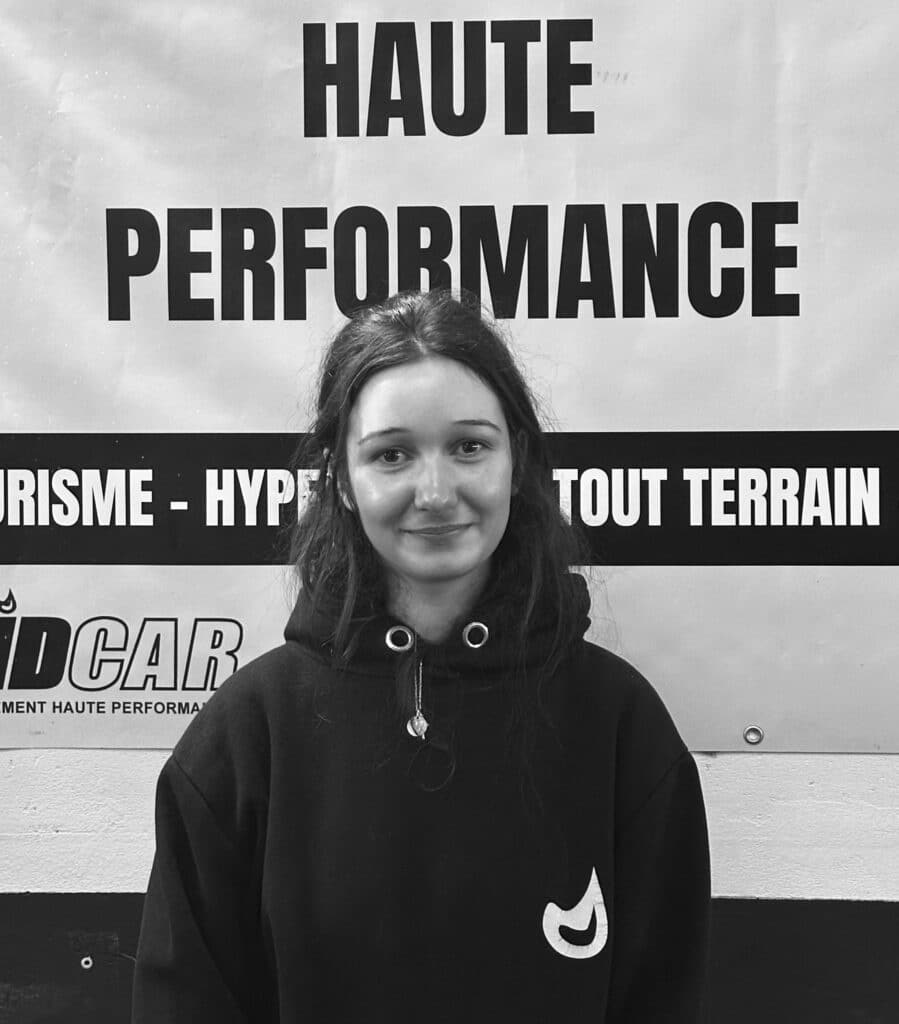 Garage Haute Performance : Jeune femme devant affiche Jeune femme souriante vêtue d'un sweat à capuche noir devant une affiche blanche et noire "VOTRE GARAGE HAUTE PERFORMANCE".