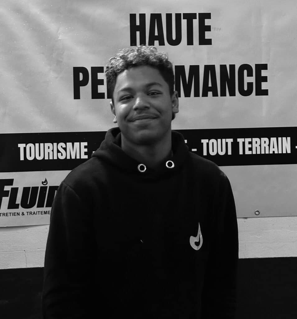 Performance et tourisme : jeune homme souriant Jeune homme souriant devant une bannière indiquant "HAUTE PERFORMANCE", "TOUT TERRAIN", et "TOURISME". Il porte un sweat à capuche noir.